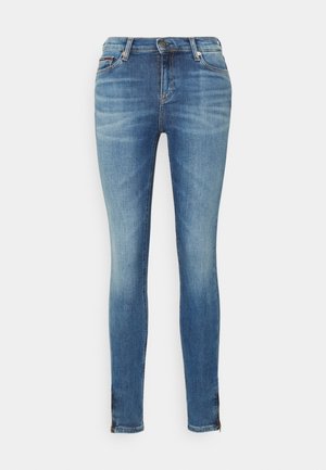 Jean skinny bleu avec légère décoloration sur les cuisses, fermeture devant par bouton et fermeture éclair, et ourlets zippés sur fond blanc.