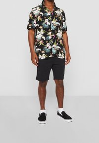 Hombre vestido con una camisa de botones de manga corta negra con flores, pantalones cortos negros, calcetines blancos y zapatillas negras sin cordones, frente a un fondo liso.