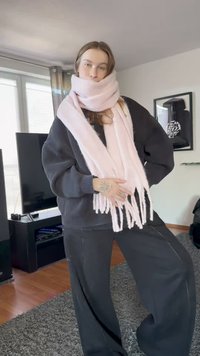 Young woman wearing oversized black sweater and pants, wrapped in a large pale pink scarf indoors, standing on a gray carpet.
