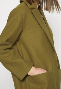 Olivgrön kappa i texturerad tyg, med spetsig krage, långa ärmar och framfickor. Skräddarsydd design med avslappnad passform.