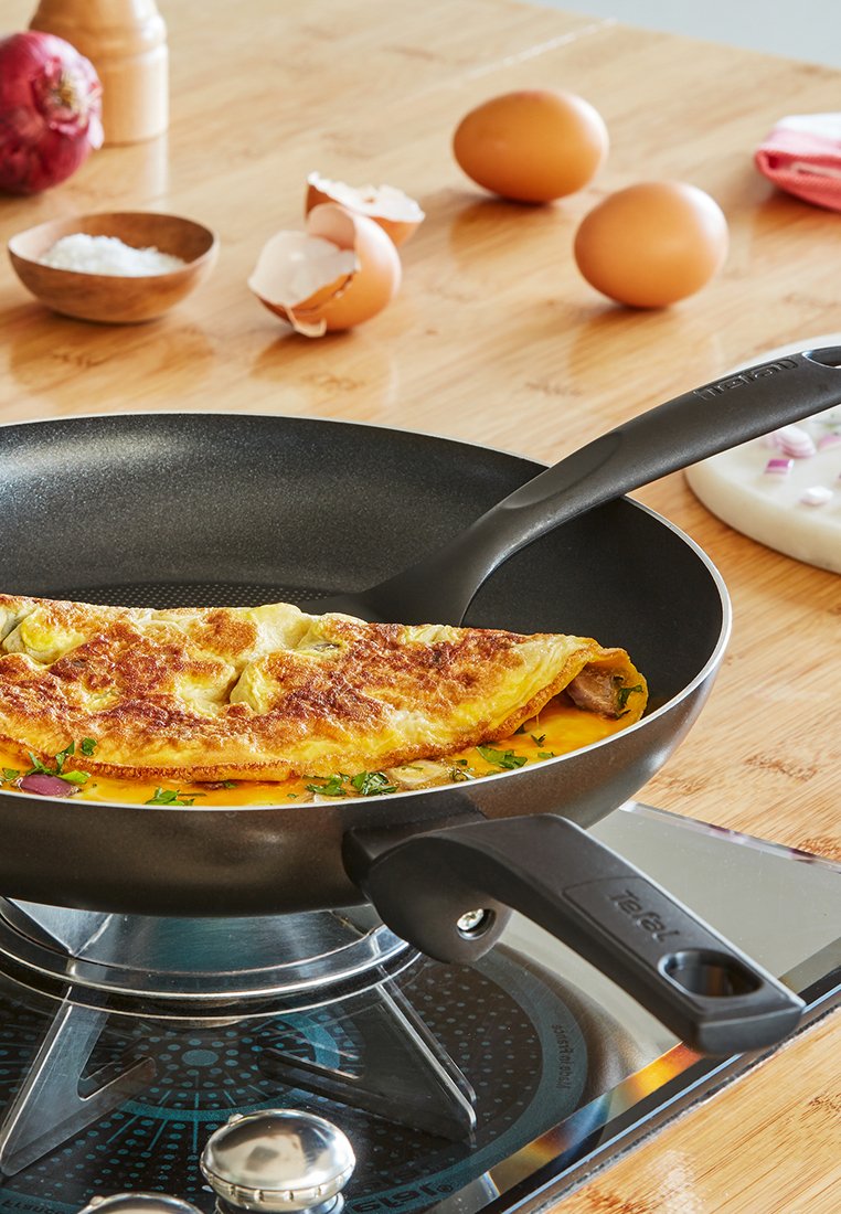 Poêle à frire antiadhésive avec une surface noire, omelette dorée et herbes vertes. Oeufs et morceaux d'oignon à proximité sur un plan de travail en bois.