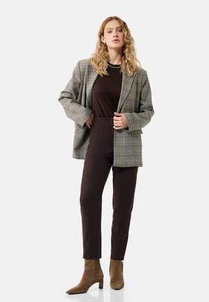 Nainen, jolla on pitkät vaaleat kiharat hiukset, pukeutuneena harmaaseen ruudulliseen bleiseriin, mustaan paitaan, mustiin housuihin ja ruskeisiin mokkanahkaisiin nilkkureihin, seisoo yksivärisen taustan edessä.