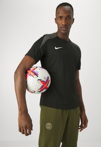 Camiseta deportiva negra de Nike con acentos grises, con cuello redondo. Sosteniendo un balón de fútbol blanco con patrones geométricos coloridos. Pantalones verde oliva.