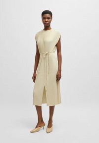 Knee-length, beige ribbed dress with short sleeves, a tie waist, and a front slit. Paired with light beige heeled mules. Smooth texture.