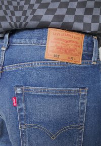 Jean en denim bleu avec des coutures orange, présentant une patch en cuir à l'arrière et un petit logo rouge sur la poche. Design classique 501.