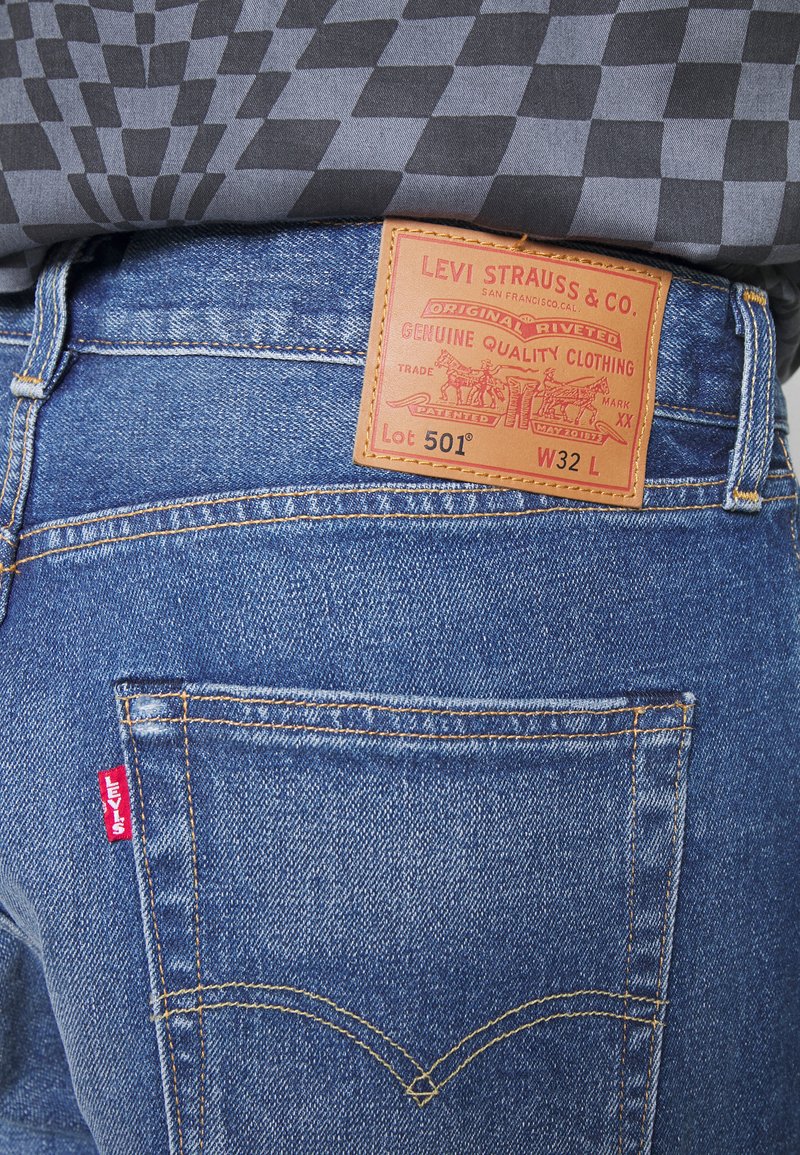 Vaqueros de mezclilla azul con costuras naranjas, con una parche de cuero en la parte trasera y un pequeño logo rojo en el bolsillo. Diseño clásico 501.