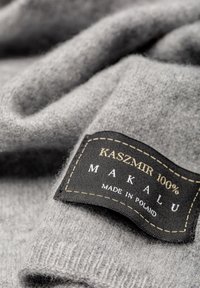 Szary materiał kaszmirowy charakteryzuje się gładką fakturą, z wyraźną czarną etykietą z napisem "KASZMIR 100% MAKALU WYPRODUKOWANE W POLSCE" z żółtym szwem.