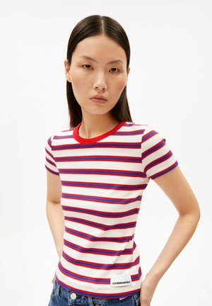 Junge Frau in einem kurzärmeligen Hemd mit weißen, roten und blauen Streifen, steht mit den Händen in der Hüfte vor einem schlichten weißen Hintergrund.