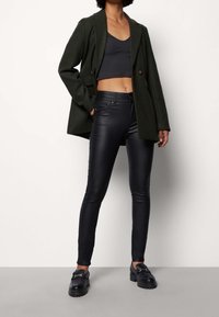 Blazer ajusté vert olive porté sur un crop top noir côtelé, associé à un pantalon skinny noir taille haute effet cuir et des mocassins noirs épais.