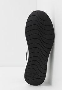 Musta kumipohja, jossa on aaltoileva urakuvio ja valkoinen reuna. Kantapäässä näkyvissä brändäys. Yleisilmeeltään sileä pinta.