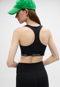 Melnas sporta krūšturis ar sacensību aizmuguri, ar Calvin Klein logotipa jostu. Valkāts kopā ar melniem augsto jostasvietu legingiem. Zaļa beisbola cepure galvā.