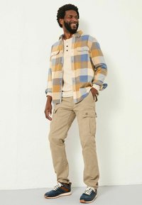 Flanellhemd in blau, gelb und weißem Karomuster, beige Cargo-Hosen und blaue Sneakers mit braunen Akzenten. Lässiges Outfit mit gestapeltem Look.