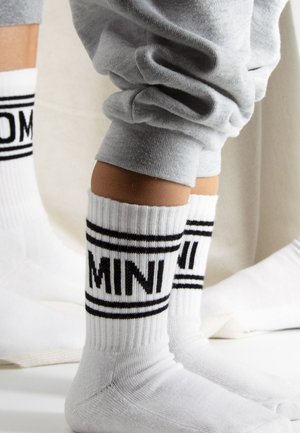 Kind staat op met witte sokken met zwarte "MINI" tekst en strepen, draagt lichtgrijze joggingbroek die boven de enkels is opgerold.