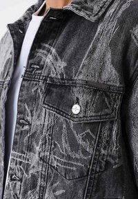 Chaqueta de mezclilla negra con detalles desgastados, botones plateados, bolsillo en el pecho y acentos de costura visibles. Tejido texturizado con patrones únicos.