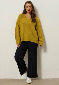 Musztardowy oversize'owy sweter dzianinowy z dekoltem w serek, zestawiony z czarnymi spodniami typu wide-leg i białymi trampkami. Miękka tekstura i luźny krój.