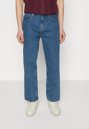 Personne portant un jean denim bleu droit, des baskets blanches et un haut sans manches texturé bordeaux, debout devant un fond uni.