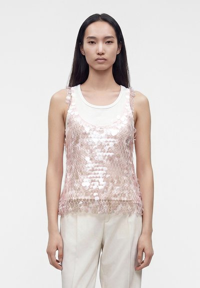 Femme aux longs cheveux noirs portant un haut blanc sans manches superposé d'un caraco en maille rose à paillettes et un pantalon blanc sur un fond uni.