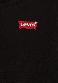 Tela negra con un logotipo de Levi's bordado en rojo en forma de insignia, con texto en blanco. La textura es suave con un ligero brillo.