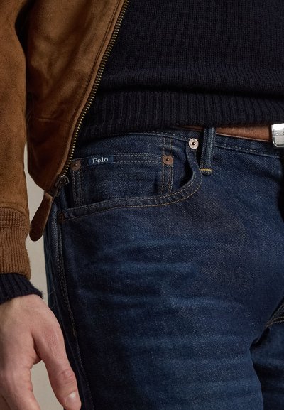 Dunkelblaue Jeans aus Denim mit subtiler Textur, ausgestattet mit kupferfarbenen Beschlägen und einem "Polo"-Etikett. Kombiniert mit einer braunen Wildlederjacke.