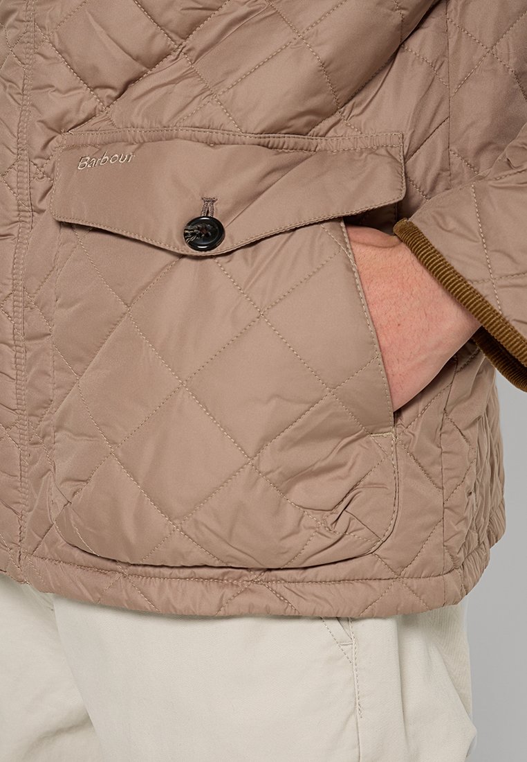 Veste matelassée beige avec une poche à rabat boutonnée, finitions en velours côtelé au niveau des poignets, et broderie discrète de la marque sur la poitrine gauche.