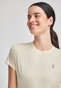 T-shirt in cotone a girocollo color beige chiaro, con una piccola figura ricamata sul lato sinistro del petto. Texture liscia, maniche corte, design aderente.