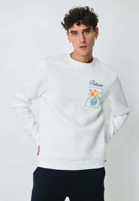 Sweatshirt blanc à col rond, doté d'une poche avec un motif floral et le texte "Palano". Le tissu semble doux avec une texture lisse.