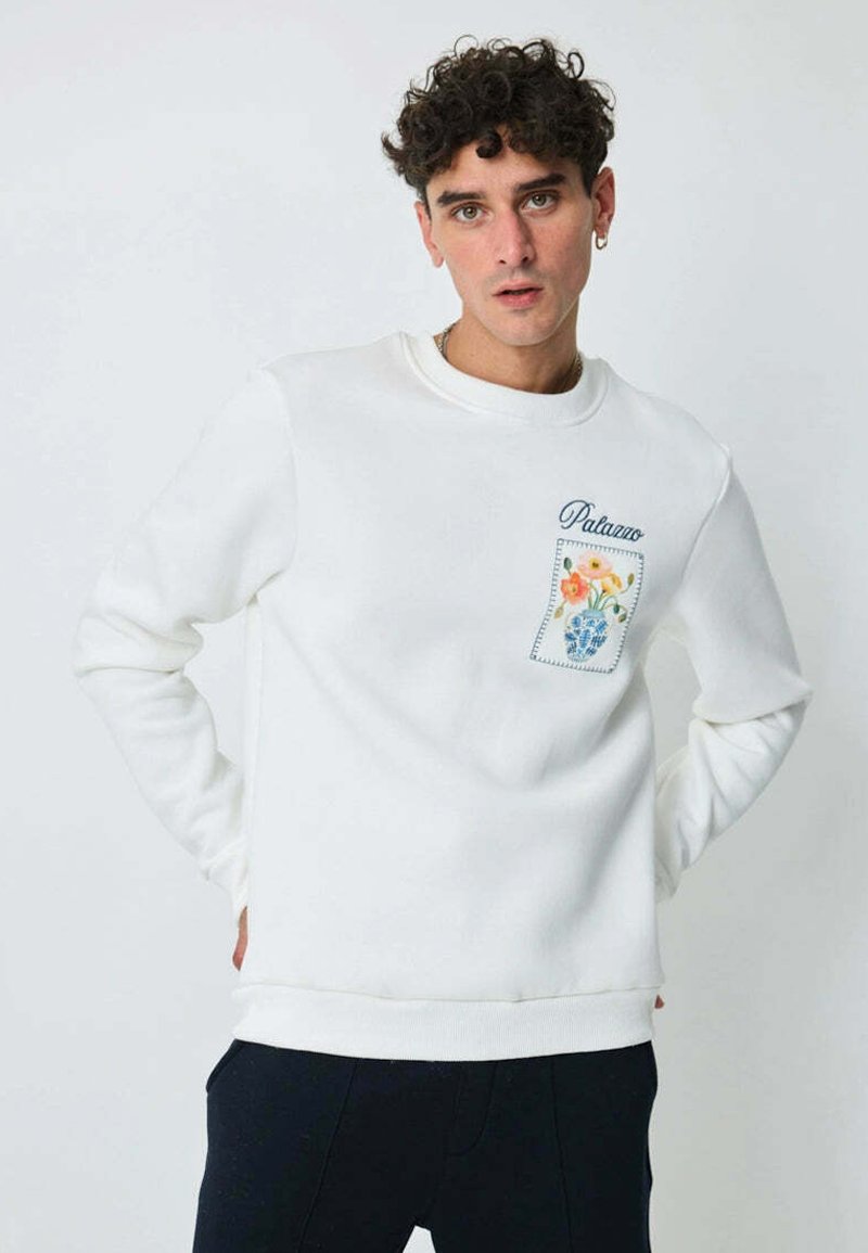 Sweatshirt blanc à col rond, doté d'une poche avec un motif floral et le texte "Palano". Le tissu semble doux avec une texture lisse.