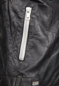 Nahaufnahme einer schwarzen Lederjacke mit einem vertikalen silbernen Reißverschluss und einem kleinen Metall-Logo mit der Aufschrift "Freaky Nation."