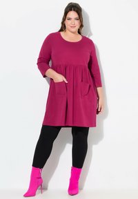 Vestido magenta de manga larga con cintura fruncida, que presenta dos bolsillos frontales. Lleva puesto con leggins negros y botines de tacón de aguja rosa brillante.