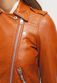 Brązowa skórzana kurtka z usztywnionym kołnierzem, srebrnym zamkiem błyskawicznym i boczną kieszenią. Charakteryzuje się gładką fakturą i szczegółami przeszyć.