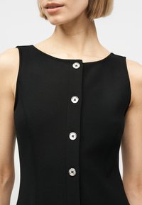 Γυναίκα που φορεί ένα μαύρο αμάνικο φόρεμα με μια σειρά από πέντε ασημένια κουμπιά στο μπροστινό μέρος και στρογγυλή λαιμόκοψη.