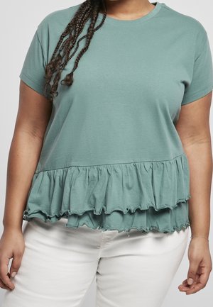 Vrouw draagt een saliegroene top met korte mouwen en een dubbel geplooide zoom, gecombineerd met een witte broek tegen een effen achtergrond.
