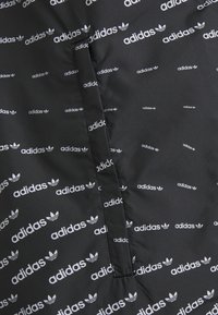 Černá tkanina s opakujícím se bílým logem Adidas a trojlístkem. Hladká textura, na levé straně je viditelný šev.