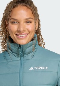 Donna sorridente con capelli ricci, indossa una giacca teal con colletto alto e logo "TERREX", su uno sfondo grigio chiaro.