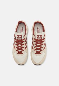 Zapatillas Adidas con parte superior en color crema, acentos rojos y tres rayas. Presenta una puntera de malla texturizada y cordones redondos. Logo en el talón.