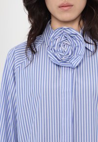 Camicia a righe blu e bianche con un ampio colletto a forma di fiore. Il tessuto appare liscio con una leggera lucentezza, accentuando il design.