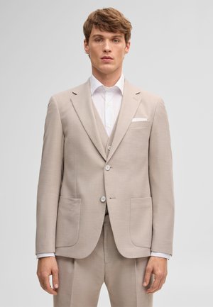 Junger Mann mit hellbraunen Haaren, der einen beigen Anzug, weißes Hemd und Weste trägt, steht vor einem schlichten hellgrauen Hintergrund.