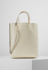 Krämfärgad läder tote-väska med korta handtag och en avtagbar axelrem i silverkedja, som står på en vit pelare.