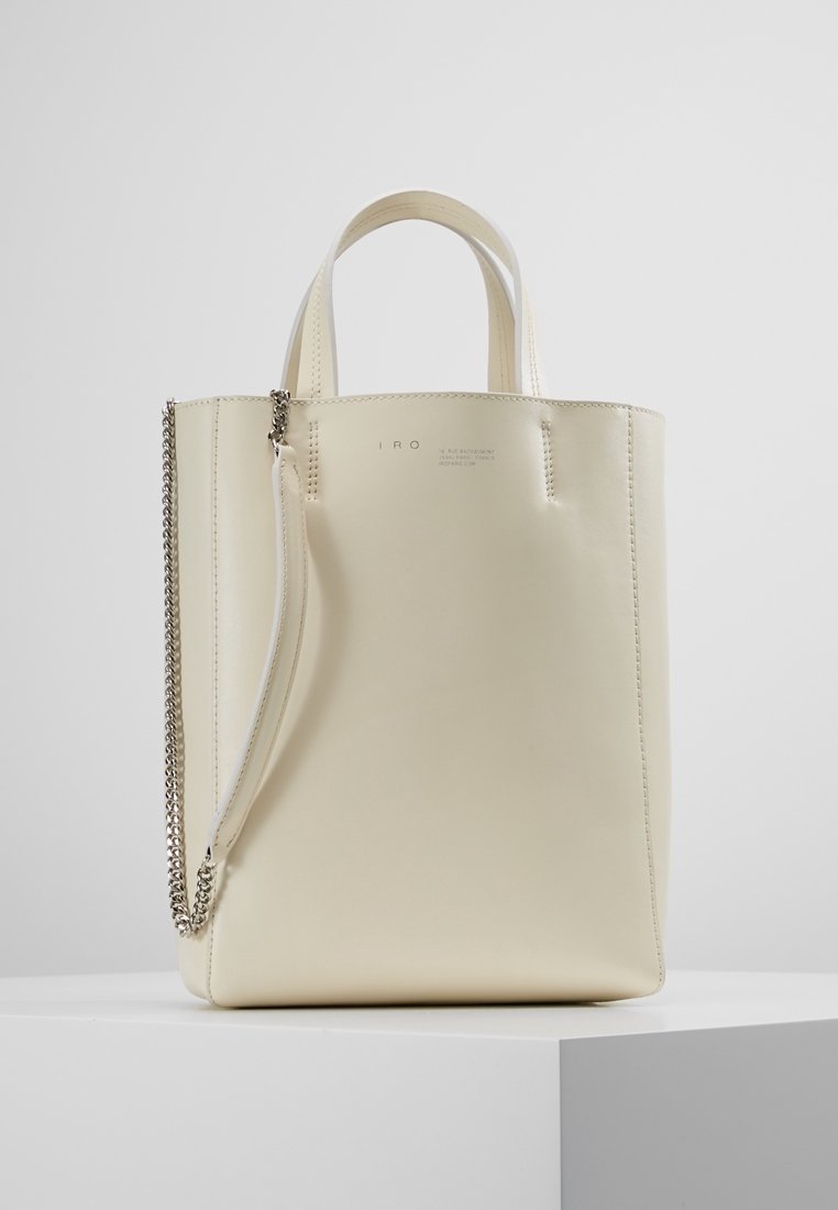 Krämfärgad läder tote-väska med korta handtag och en avtagbar axelrem i silverkedja, som står på en vit pelare.