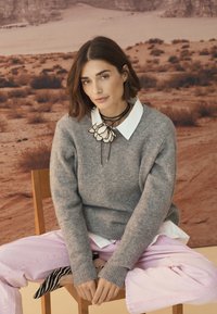 Sivo volneno pulover z rebrastimi rokavi, pod njim bela srajca z ovratnikom, roza jeans hlače. Cvečkasta choker ogrlica, sedi na leseni stolu.