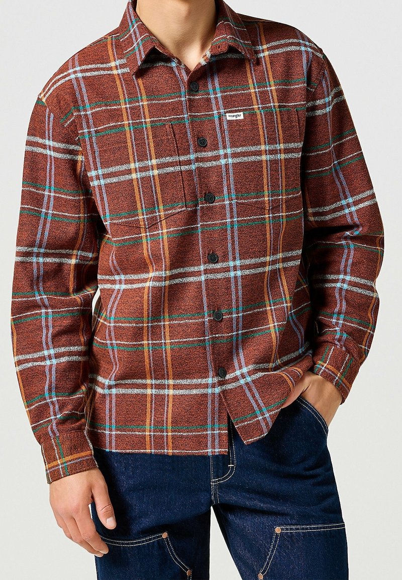 Homme portant une chemise à carreaux marron, orange et bleue avec une poche poitrine, assortie à un pantalon en jean bleu foncé.