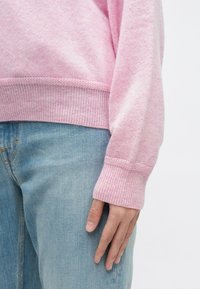 Pulover roz deschis dintr-un material tricotat moale, cu manșete și tiv cu ajur, purtat împreună cu blugi denim albaștri deschiși.