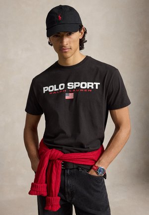 Ung mand iført en sort Polo Sport t-shirt, sort kasket, rød sweater bundet om livet, sorte jeans og et blåt ur på håndleddet.
