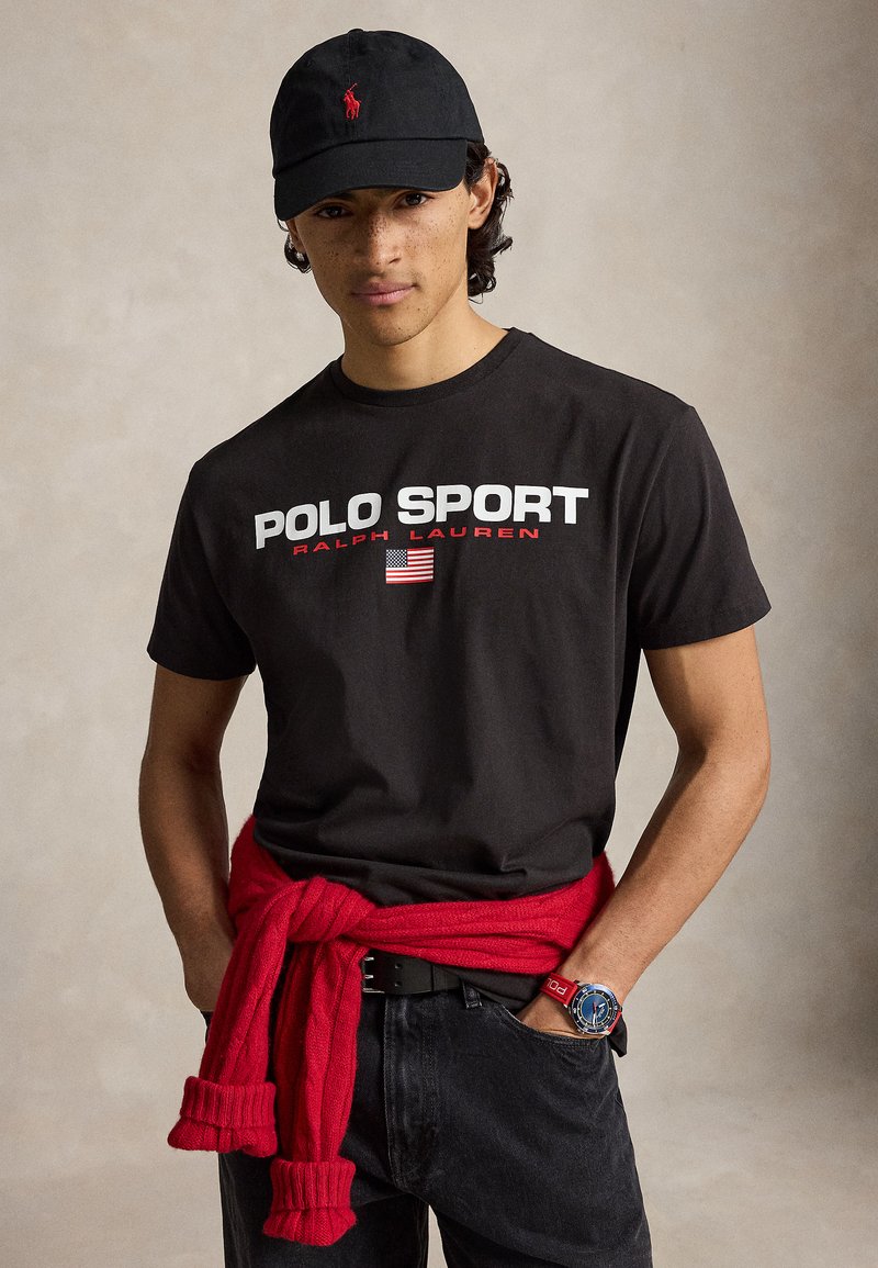 Νέος άνδρας με μαύρο μπλουζάκι Polo Sport, μαύρο καπέλο, κόκκινο πουλόβερ δεμένο γύρω από τη μέση, μαύρα τζιν και μπλε ρολόι στον καρπό.