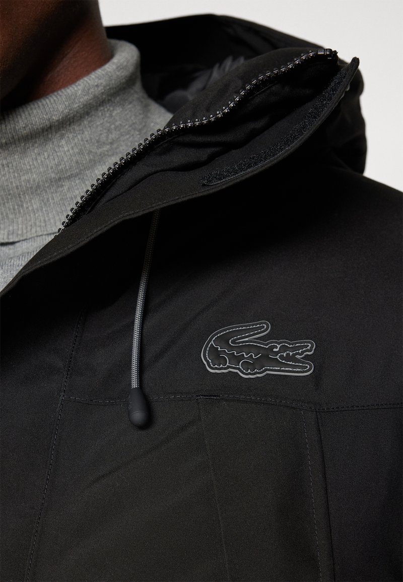 Chaqueta negra con cuello alto, que presenta un logo de cocodrilo negro de marca. Tejido resistente al agua con textura suave y cierre frontal de cremallera.