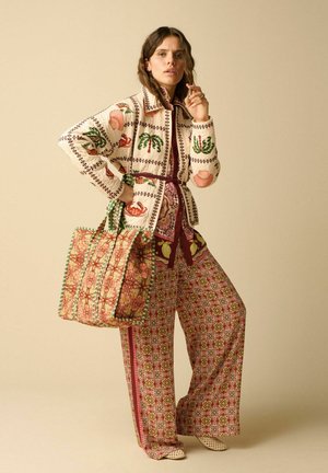 Woman wearing a patterned jacket and wide-leg pants, holding a large floral tote bag, standing against a beige background.