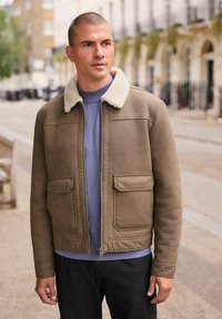 Braune Kunstlederjacke mit Schaffellkragen, zwei vorderen Taschen und Reißverschluss, getragen über einem blauen Hemd und dunklen Hosen.