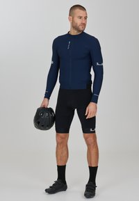 Jersey da ciclismo a maniche lunghe blu navy con zip frontale, abbinato a pantaloni corti neri e calze. La persona tiene un casco da ciclismo nero.