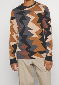 Koszula z długim rękawem z wzorem w zygzaku w odcieniach brązu, czerni i kremu, z okrągłym dekoltem oraz małym białym logo na piersi.