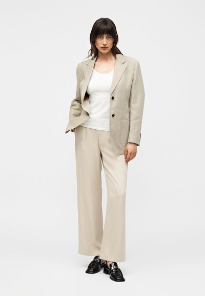 Γυναίκα στέκεται σε λευκό φόντο, φοράει μπεζ σακάκι, ταιριαστά φαρδιά παντελόνια, λευκό τοπ και μαύρα μοκασίνια, με το ένα χέρι στην τσέπη.